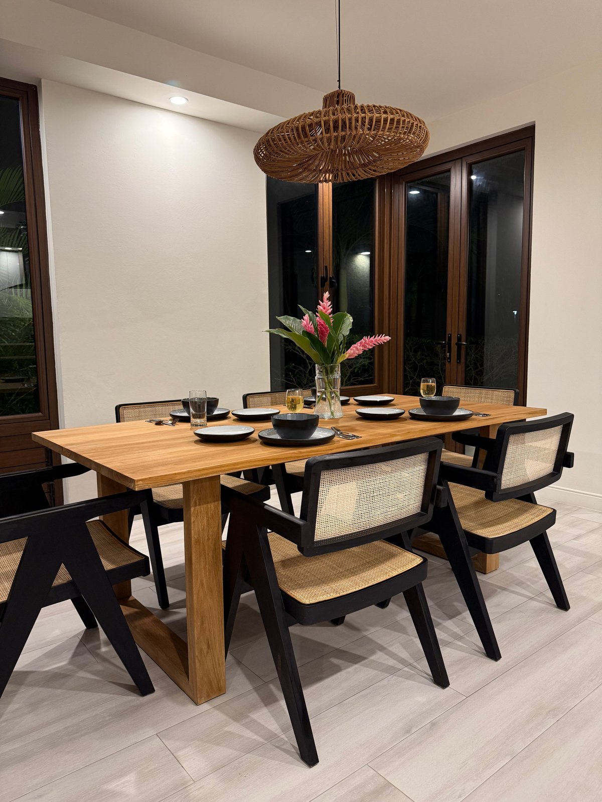 Modern dining room with wooden table, black and rattan chairs, woven pendant light, and pink flowers in a vase