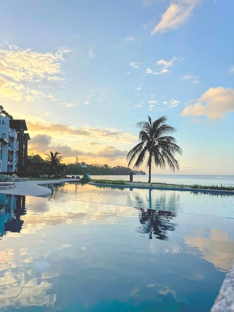 Serene tropical sunset reflected in still pool water, with palm tree and beachfront resort building