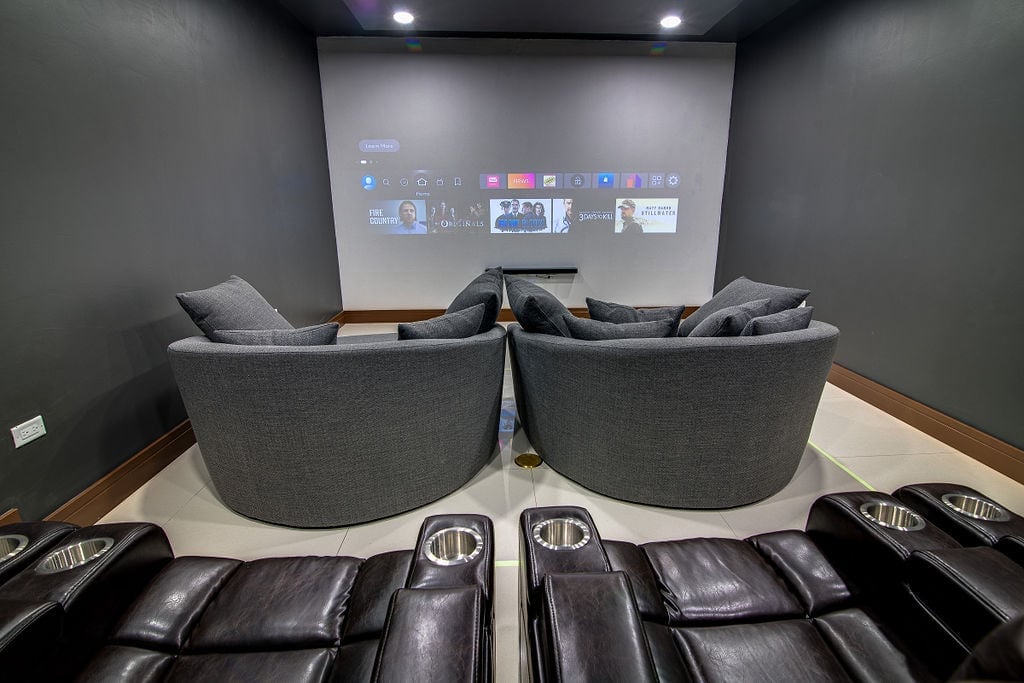 Home theater room with two gray armchairs facing a projection screen displaying media content, with black leather recliners in foreground
