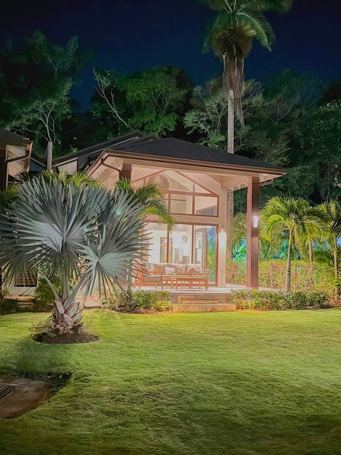 Modern garden pavilion with tan wood exterior, large windows, and black roof, surrounded by tropical plants and manicured lawn at night