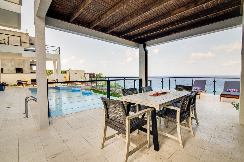 Seaside terrace with dining table, swimming pool, and ocean view under wooden pergola