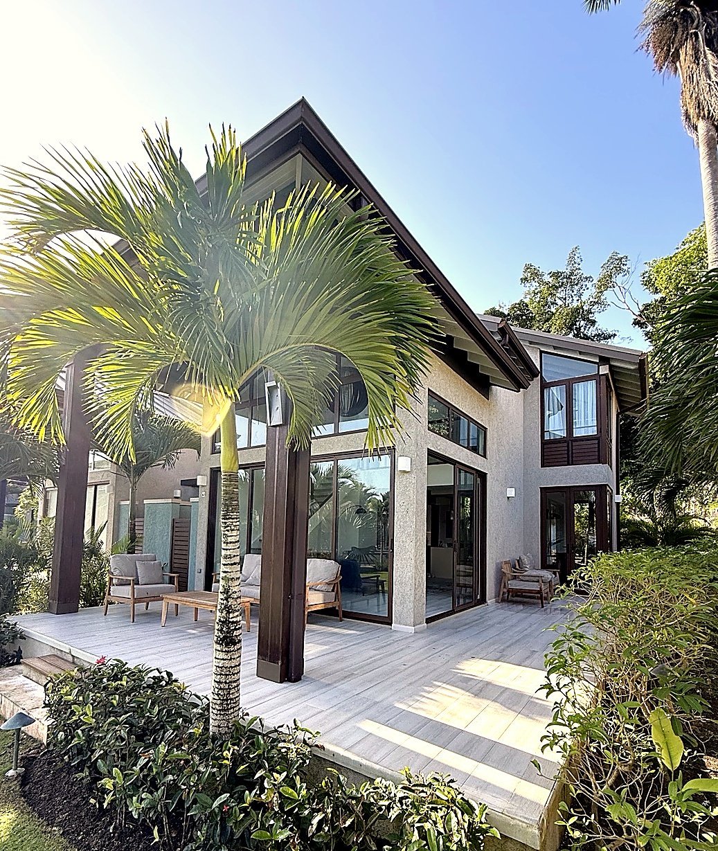 Modern home with dark roof and glass doors, surrounded by palm trees and tropical landscaping, featuring a spacious patio area